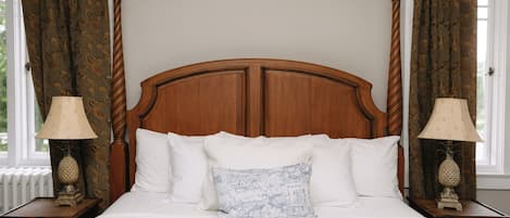 A bed with a wooden headboard, white bedding, and a decorative pillow. There are two nightstands with lamps and a window with curtains.