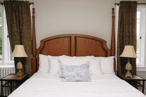 Una cama con cabecera de madera, ropa de cama blanca y una almohada decorativa. Hay dos mesitas de noche con lámparas y una ventana con cortinas.