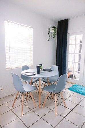 A dining area with a round table, four chairs, a potted plant, and a window with blinds. - Midcentury Style Furnished 2B/1B House Near Hospitals, Downtown & Airport (Tampa)
