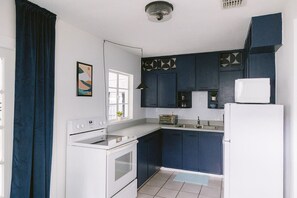 A compact kitchen with a stove, sink, and refrigerator, featuring dark blue cabinets and a white countertop. - Midcentury Style Furnished 2B/1B House Near Hospitals, Downtown & Airport (Tampa)