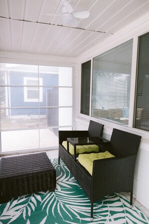 A room with a ceiling fan, a large window with blinds, a wicker sofa with cushions, and a patterned rug. - Midcentury Style Furnished 2B/1B House Near Hospitals, Downtown & Airport (Tampa)