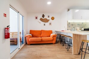 Un espace de vie compact comprenant un canapé, un coin repas avec des tabourets, une cuisine avec un plan de travail et un extincteur fixé au mur.
