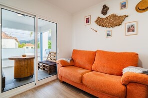 Een moderne woonkamer met een grote oranje bank, een houten salontafel en uitzicht door een glazen deur op een buitenruimte.