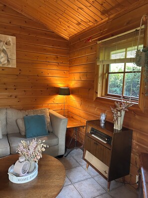 L’intérieur d’un chalet accueillant, avec un revêtement mural en bois, un canapé, une table agrémentée d’un objet décoratif, une lampe et une fenêtre équipée de stores.