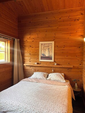 Une chambre dans une cabane en bois, avec un lit, un tableau encadré au mur et une petite table sur laquelle est posé un vase.