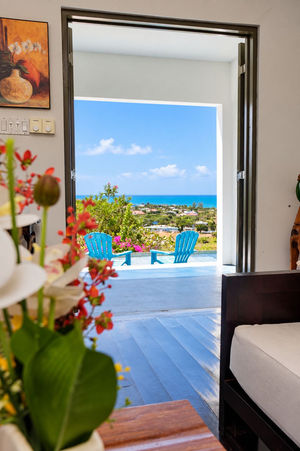 An open doorway leading to a balcony with a view of the ocean and a town. There are two blue chairs on the balcony.