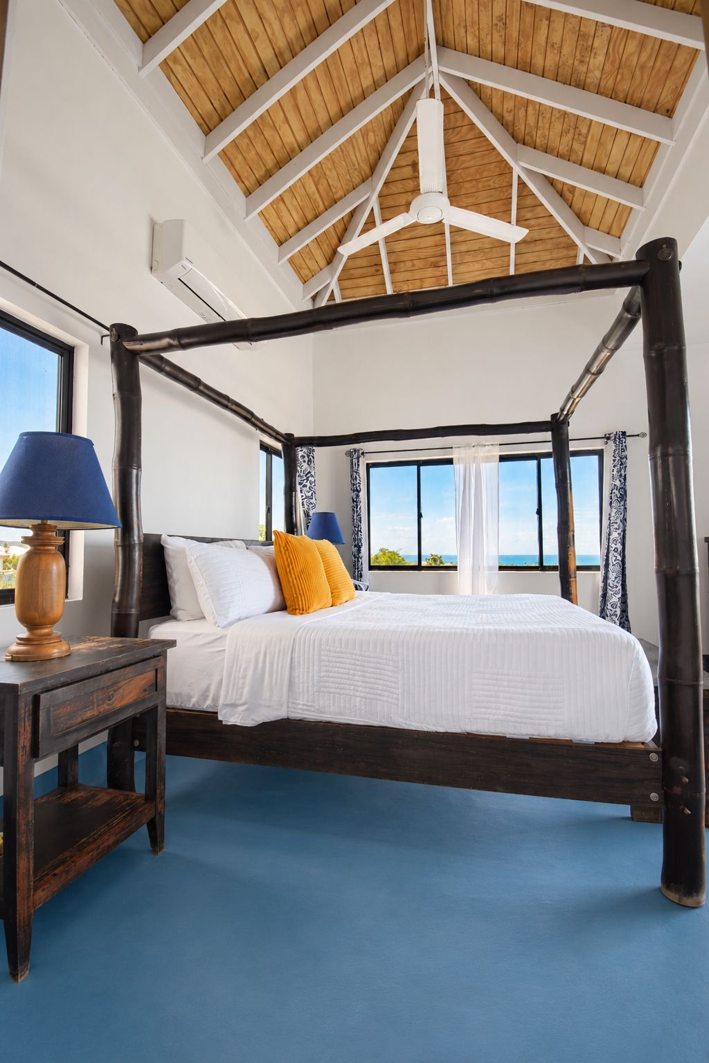 A four-poster bed with a white bedspread and two pillows, a wooden nightstand with a lamp, and a view of the ocean through large windows.