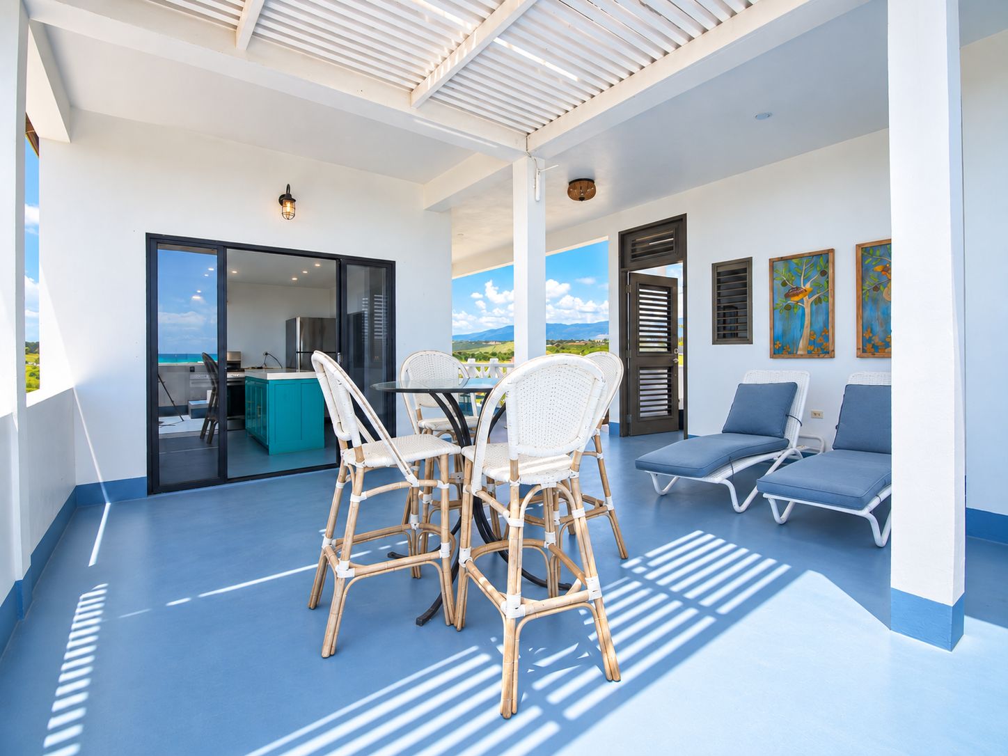 A covered outdoor area with a dining table and chairs, a kitchen visible through a glass door, and a seating area with lounge chairs.