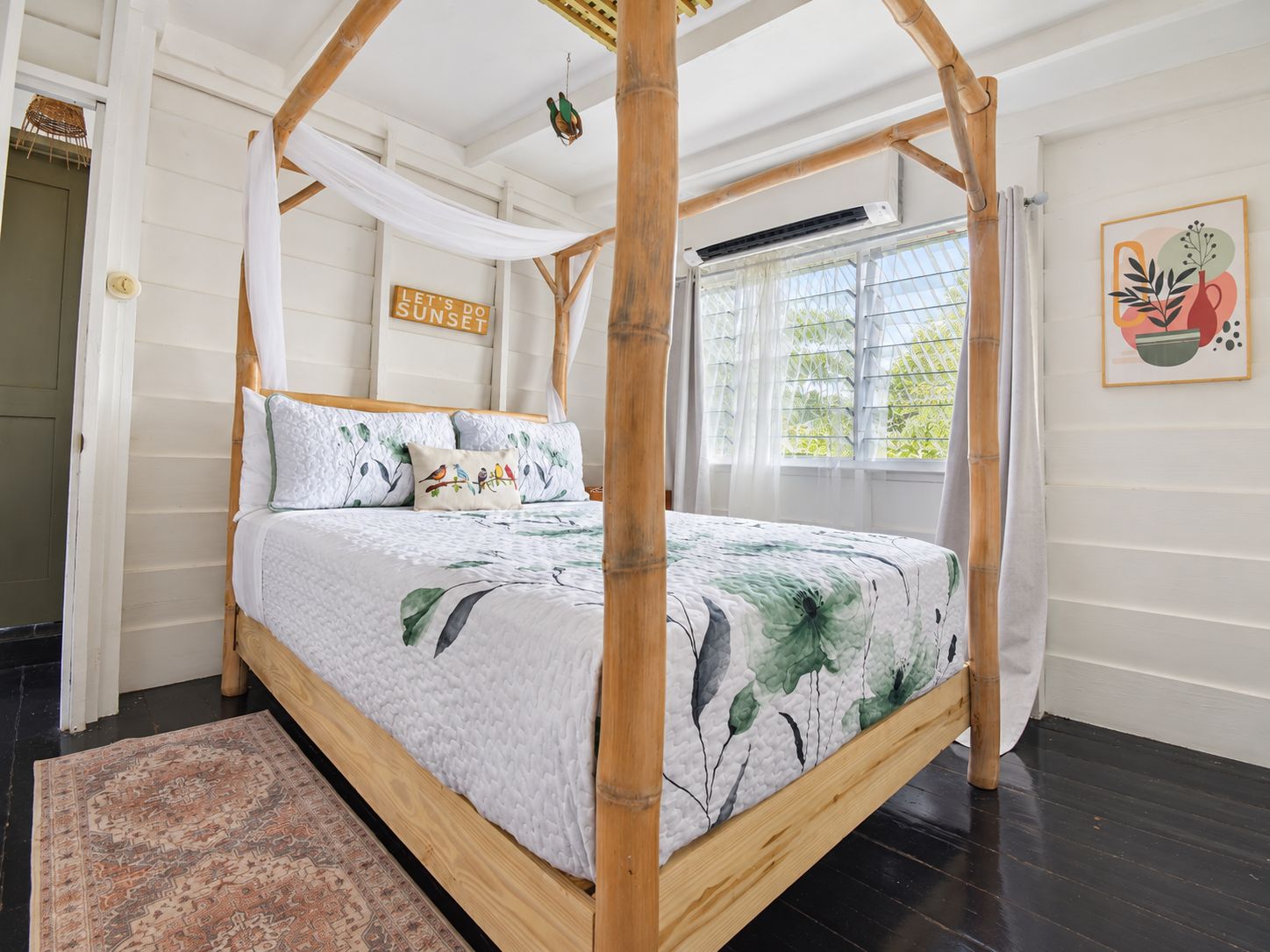 A four-poster bed with a floral bedspread, a 'Let's do sunset' sign, and a window with curtains.