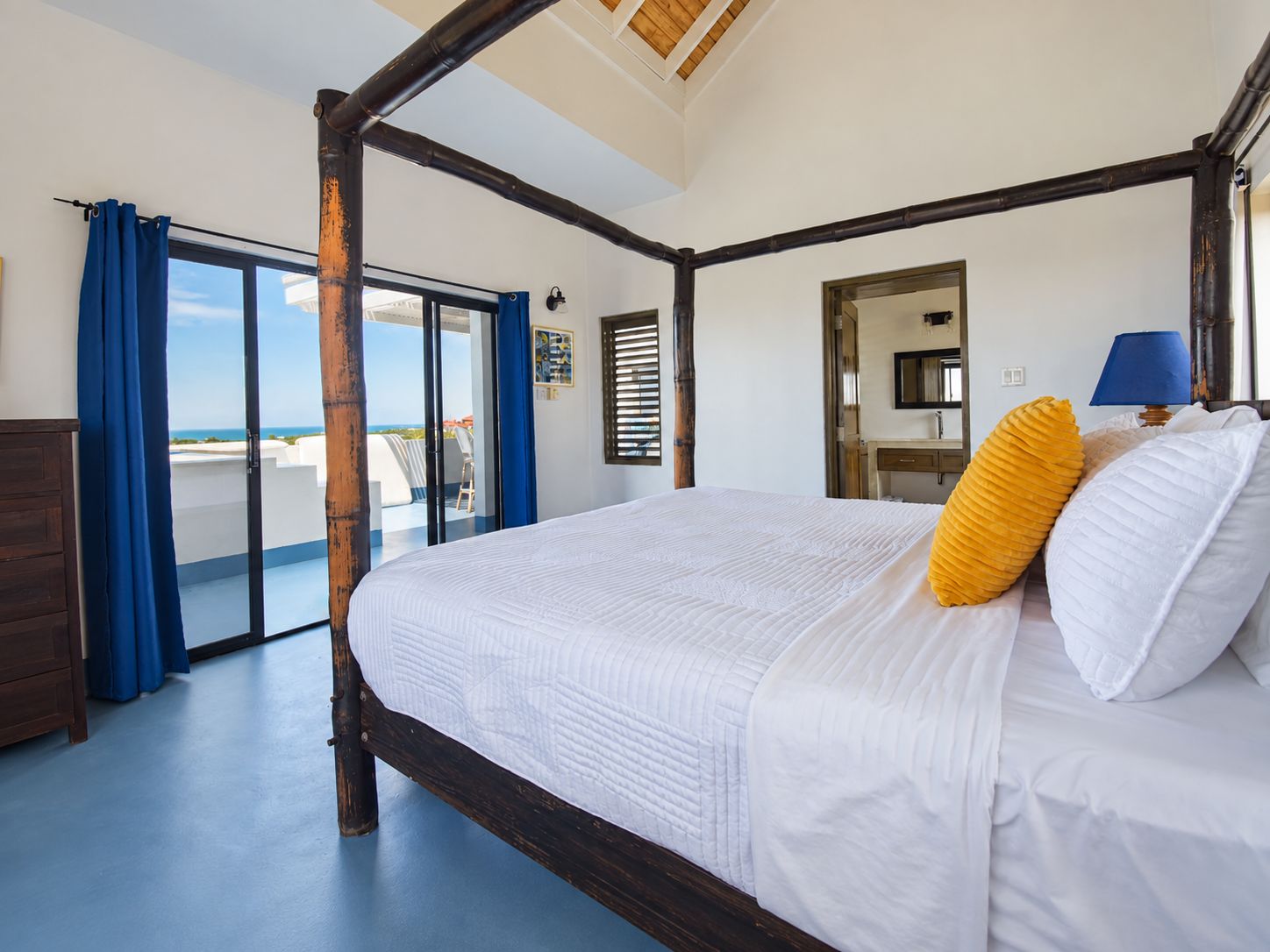 Una cama con dosel, ropa de cama blanca y dos almohadas, suelo de madera, un balcón con cortinas azules y vista al mar.