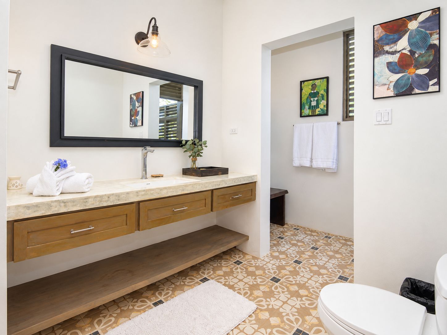 A bathroom with a large mirror, a sink with a marble countertop, wooden cabinets, a white toilet, patterned floor tiles, and a small plant on the counter.