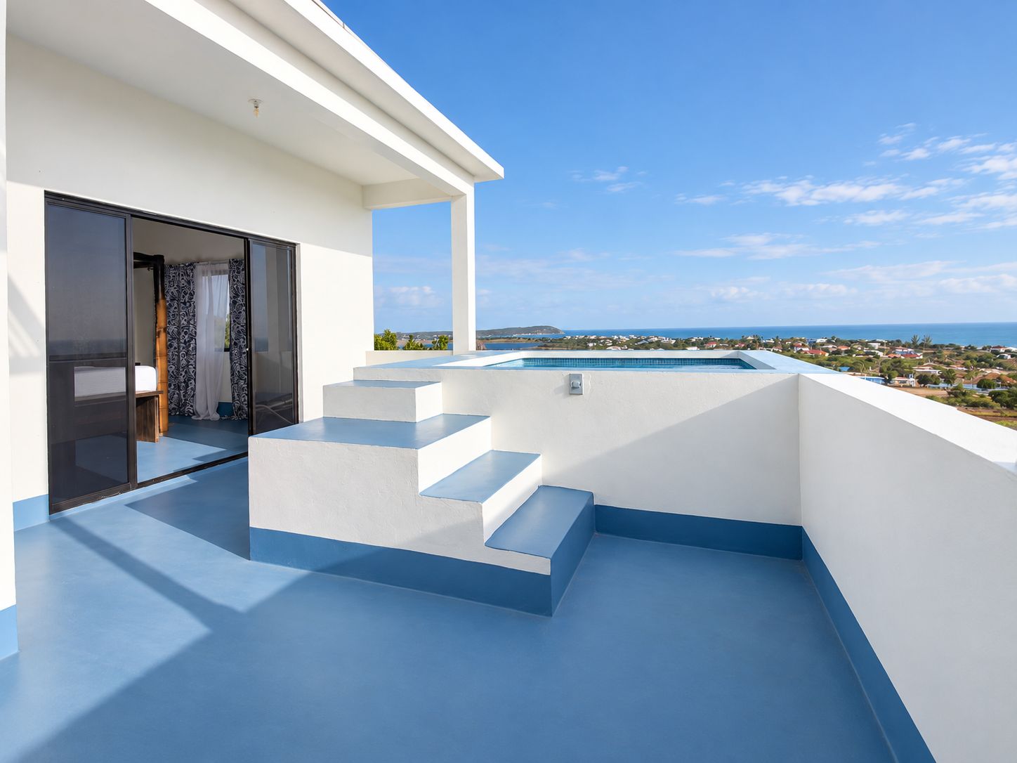 Un área de piscina moderna al aire libre con escalones que conducen a un puerta de vidrio, con vistas a un paisaje costero.