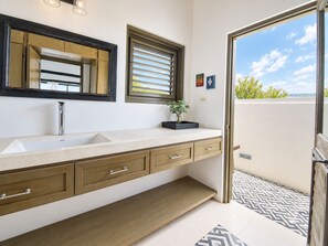 A bathroom with a large mirror, a sink, a window with blinds, and a small plant on the counter. - Villa Azure (Treasure Beach)