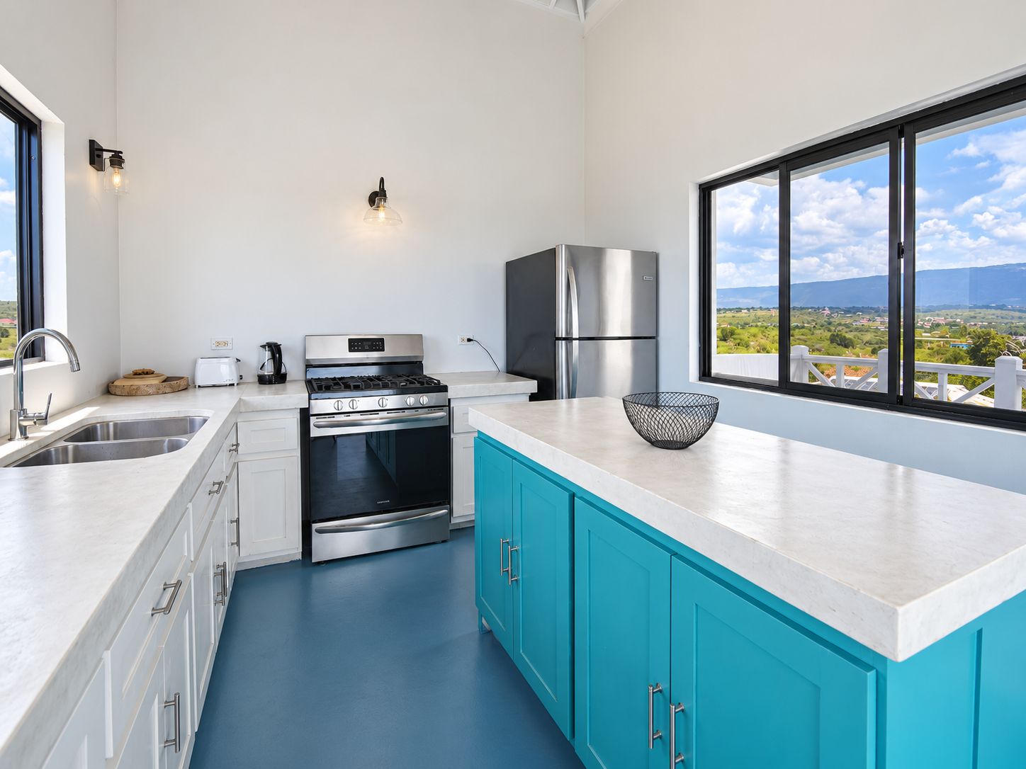 A modern kitchen with a stainless steel stove, oven, and refrigerator, a large window with a view, and a blue countertop.