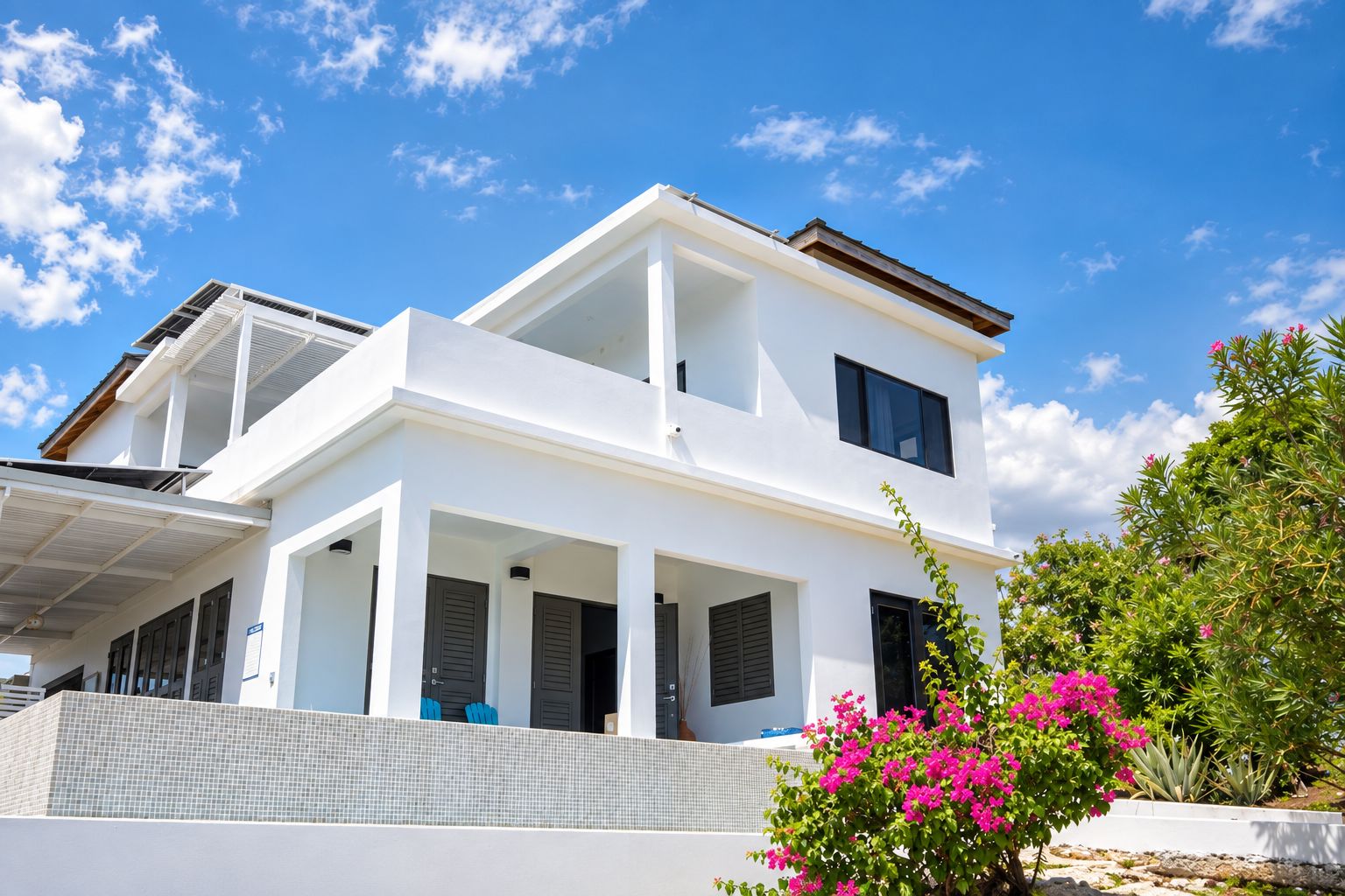 A two-story white house with a balcony, a covered patio, and a garden with pink flowers.