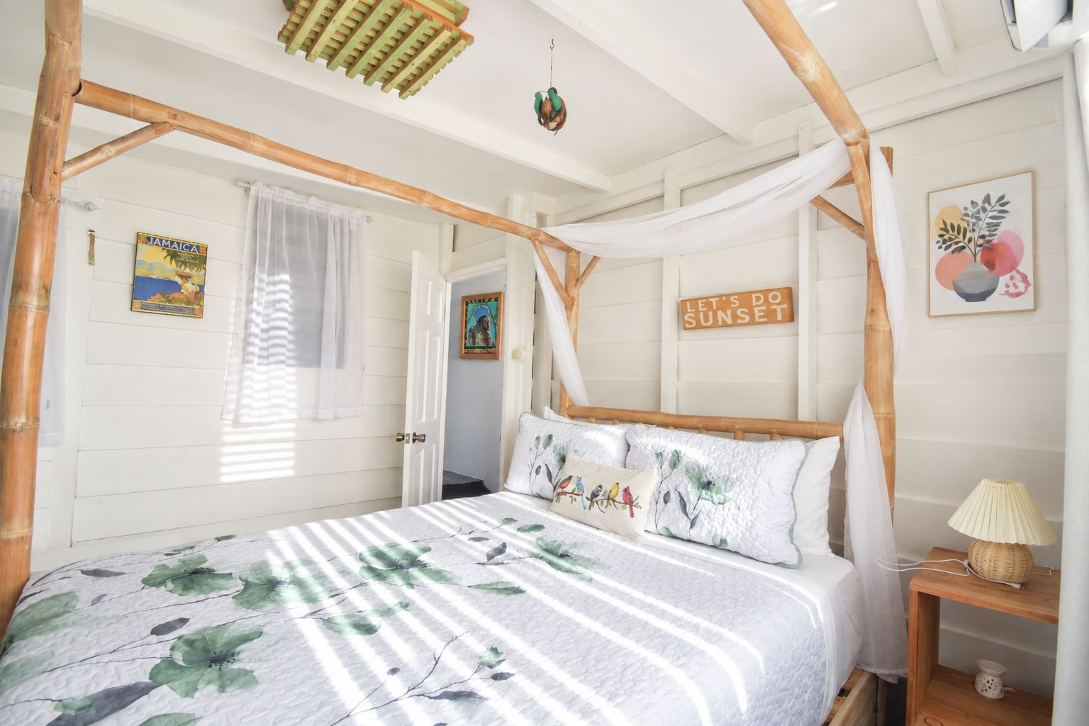 A room with a bed under a bamboo canopy, a wall with a 'Let's Do Sunset' sign, and a framed picture of birds.