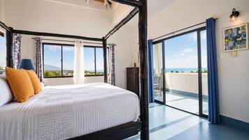 A four-poster bed with a view of the sea, a balcony with a chair, and a painting on the wall.