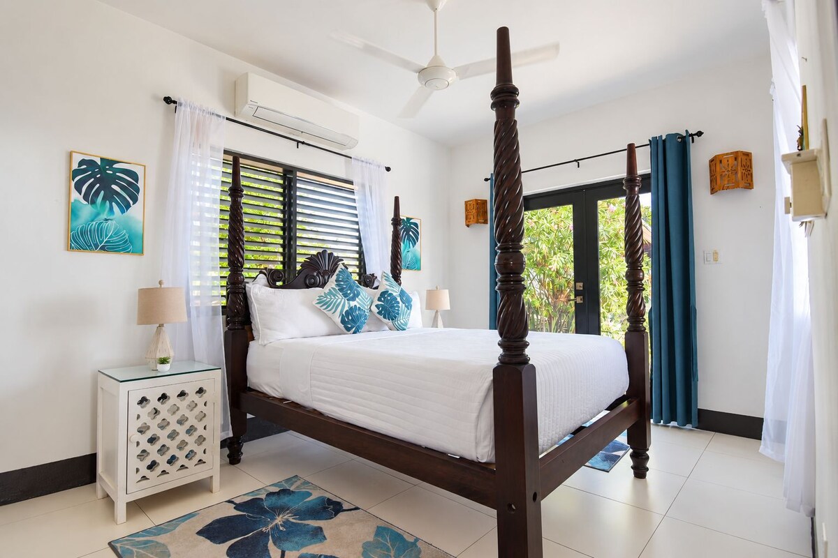 A four-poster bed with a wooden frame, white bedding, and blue and white patterned pillows. There is a white nightstand with a lamp, a blue floral rug, and a window with blinds.