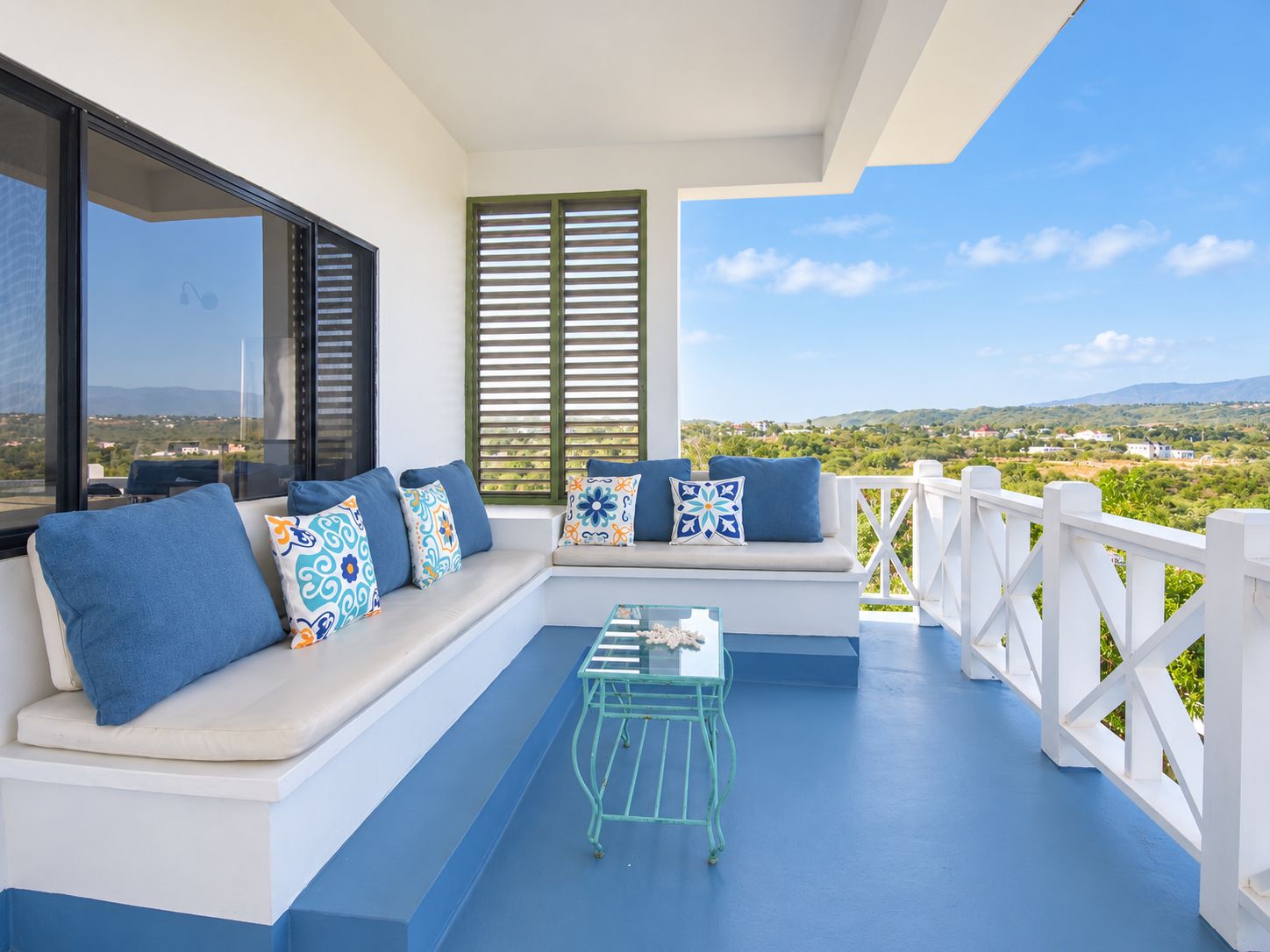 Een balkon met een witte railing, een witte bank met blauwe kussens en een glazen salontafel.
