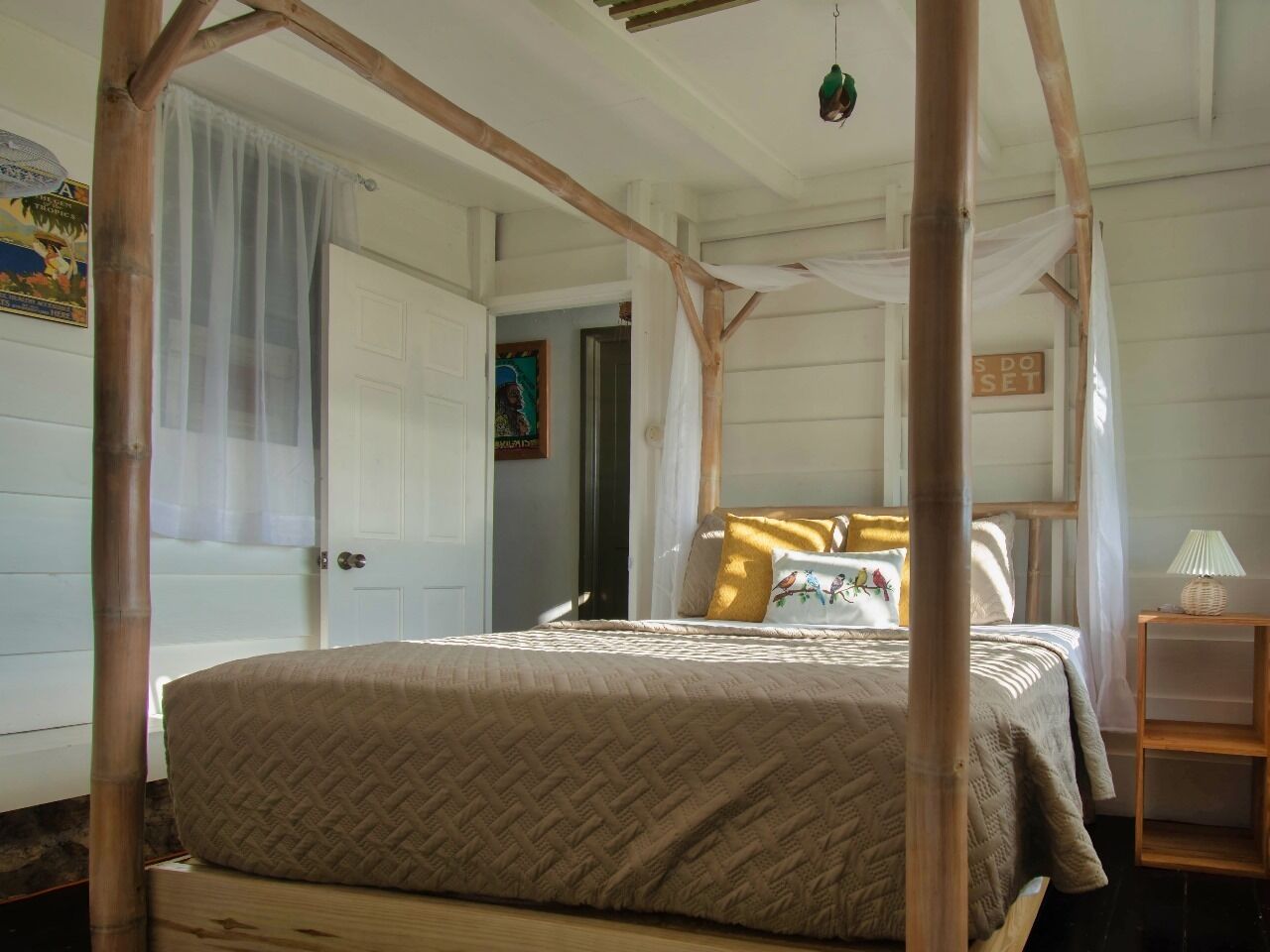 A room with a bed under a wooden canopy, a bedside table with a lamp, and a door with a curtain.