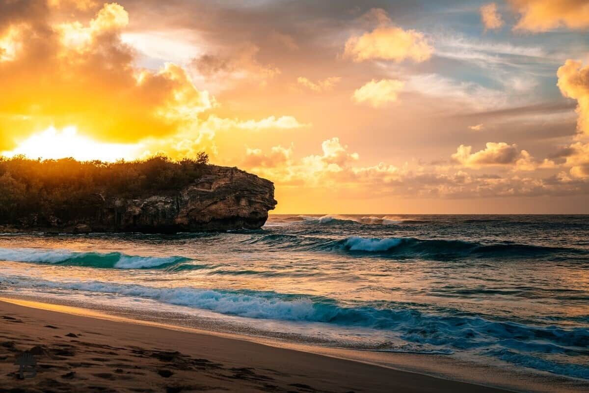 Une plage avec des vagues, un promontoire rocheux et un coucher de soleil.