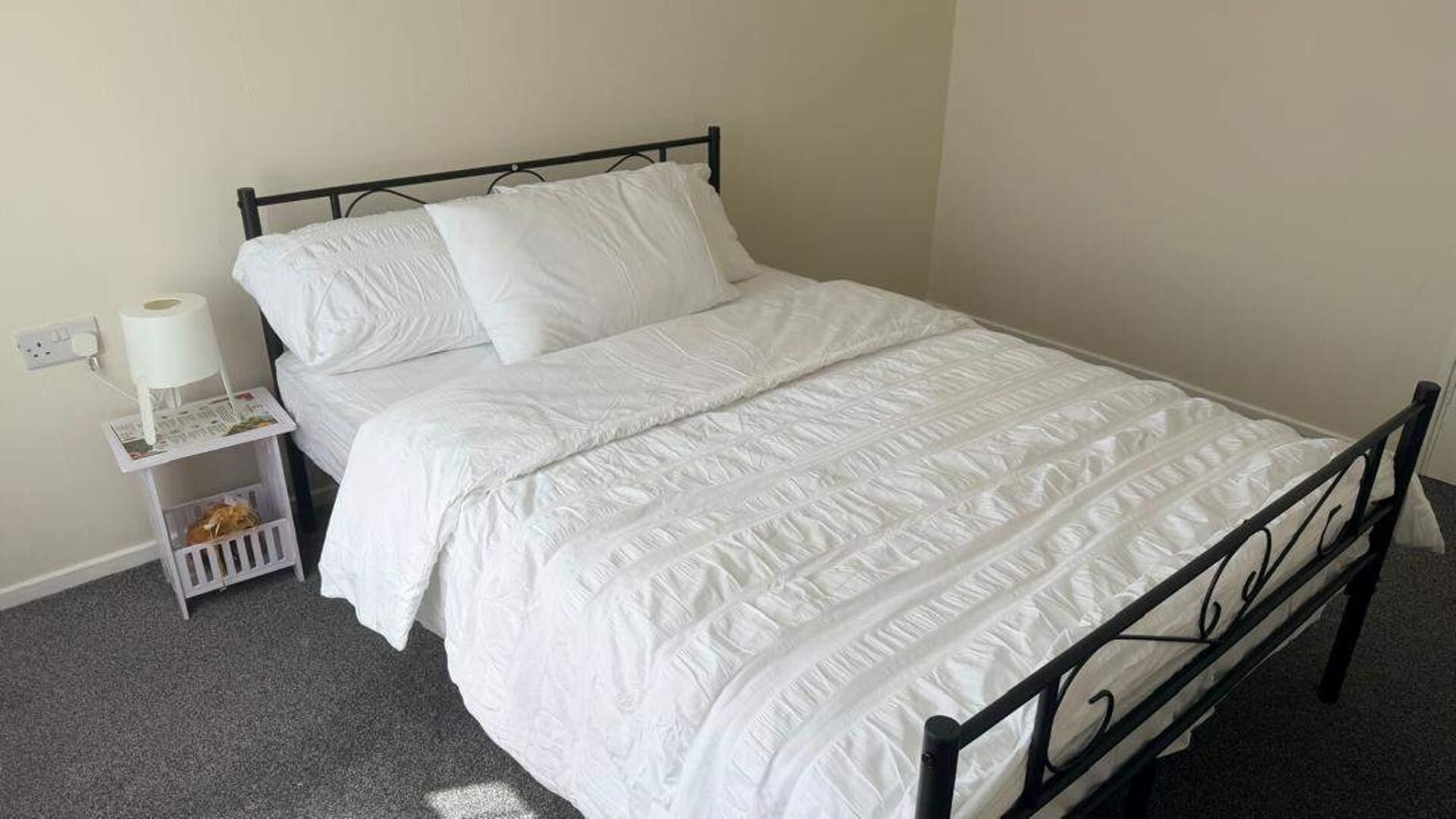Ein Einzelbett mit weißer Matratze und Kissen, ein Nachttisch mit Lampe und Zeitschrift sowie eine Steckdose an der Wand.