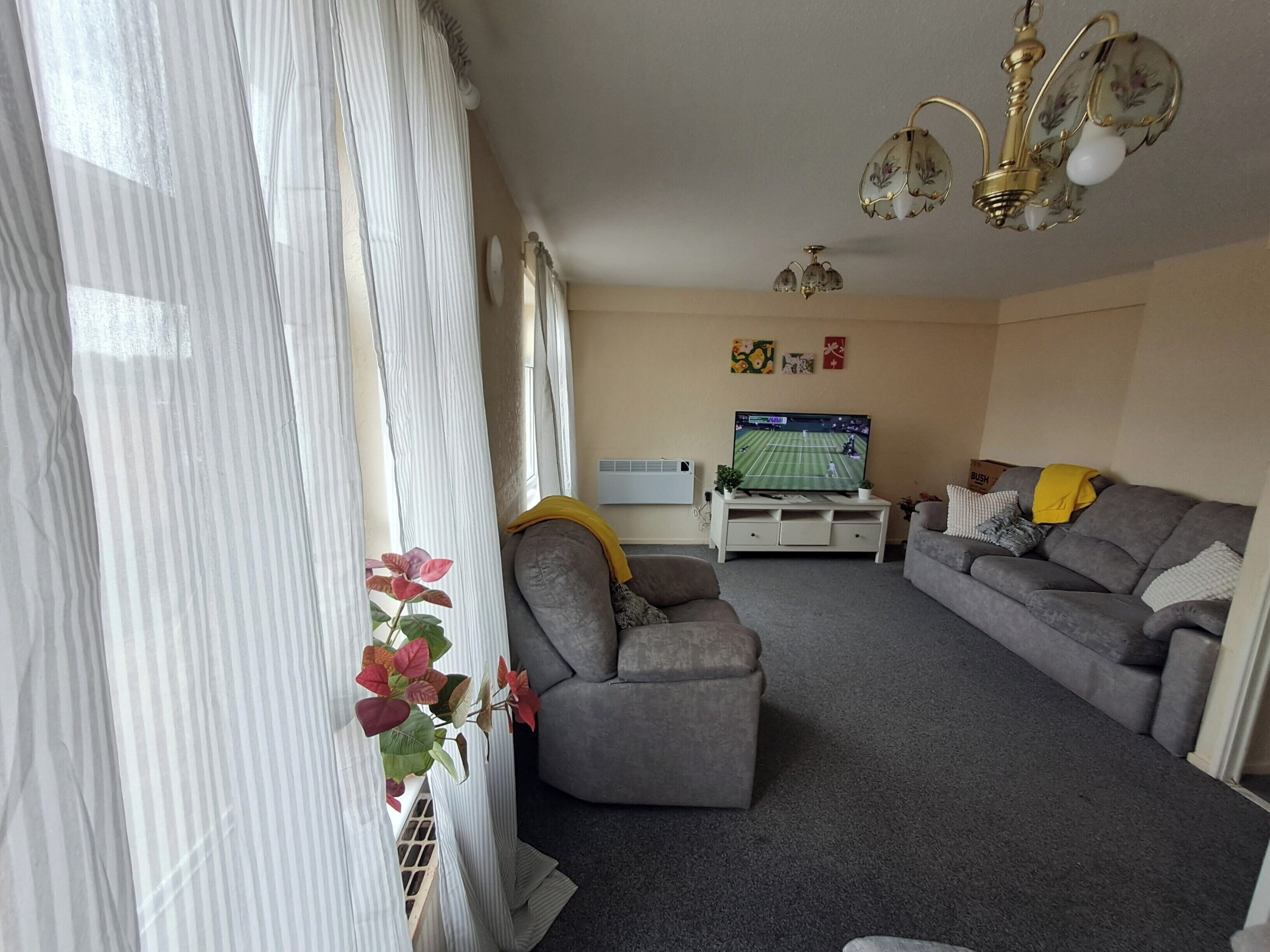 Ein Wohnzimmer mit grauem Sofa, einer weißen Unterhaltungsecke, einem Fernseher und einem Fenster mit Vorhängen.