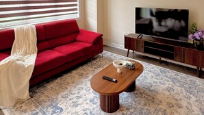 Ein Wohnzimmer mit einem roten Sofa, einem Holztisch, einem Fernseher auf einem Holzschrank und einem gemusterten Teppich.
