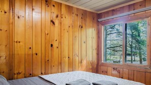 Una habitación con paredes de madera, una cama con colcha, una ventana con vista a árboles y una lámpara en el techo.