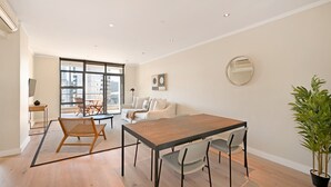 A modern living room with a dining area, large windows, and a potted plant. - Two Bed Apt | Foreshore | Pool | Gym | Secure (Cape Town)