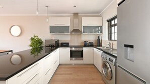 A modern kitchen with white cabinetry, a black countertop, a built-in oven, a washing machine, and a window providing natural light. - Two Bed Apt | Foreshore | Pool | Gym | Secure (Cape Town)
