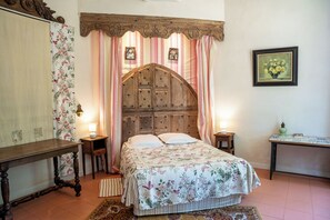 Un dormitorio con cabecera de madera, edredón floral, dos lámparas de noche, una mesita de noche, un cuadro en la pared y una pequeña mesa con un libro y una botella.