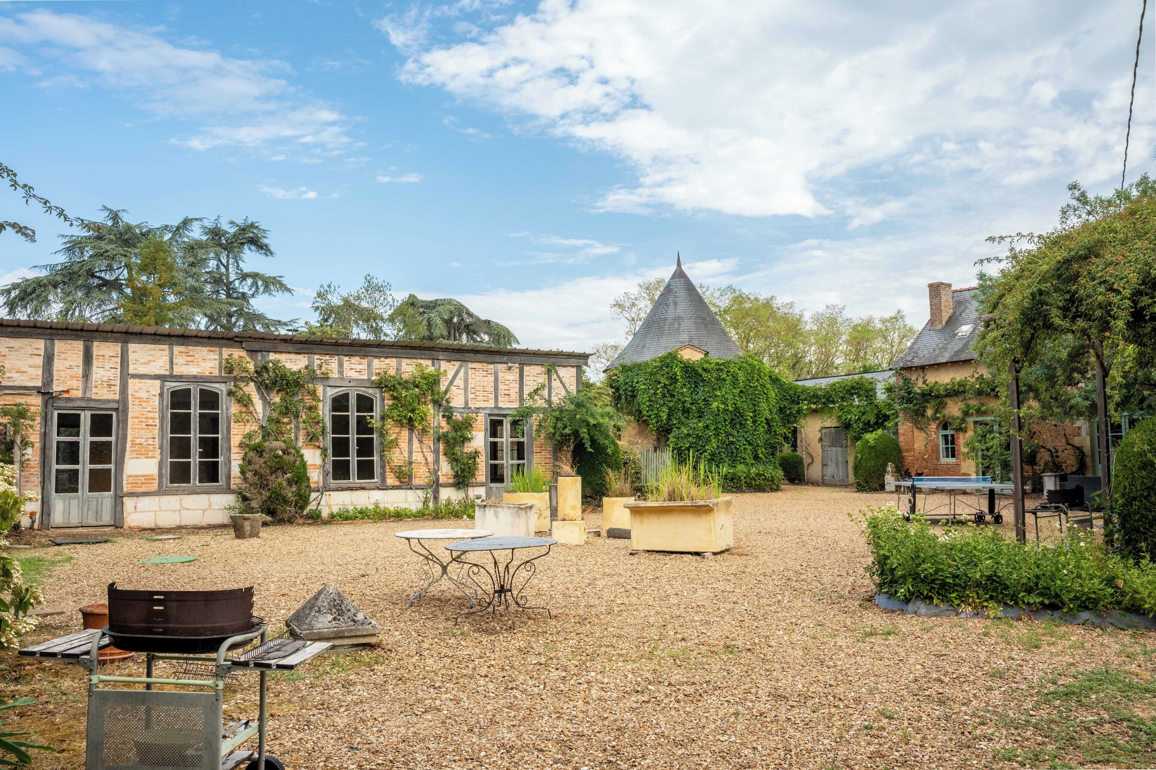 Une cour dallée de gravier, avec une table et des chaises en métal, entourée de bâtiments en briques dotés de grandes fenêtres et surmontés d’une structure de toit conique.