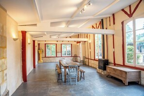 Una habitación amplia con una gran mesa de madera, bancos, chimenea y ventanas grandes.