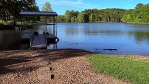 Un muelle sobre el lago con un área de descanso techada, rodeado de árboles y un cielo despejado.