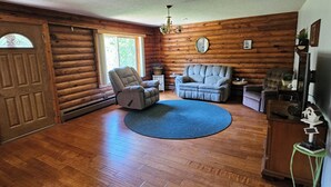 A log cabin interior with a wooden floor, a blue rug, a fireplace, and a large window.