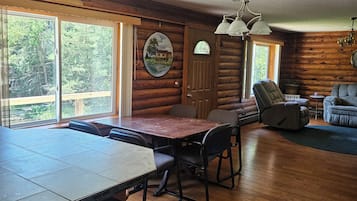 A spacious room with a wooden dining table, chairs, a sofa, and a large window offering a view of the outdoors.