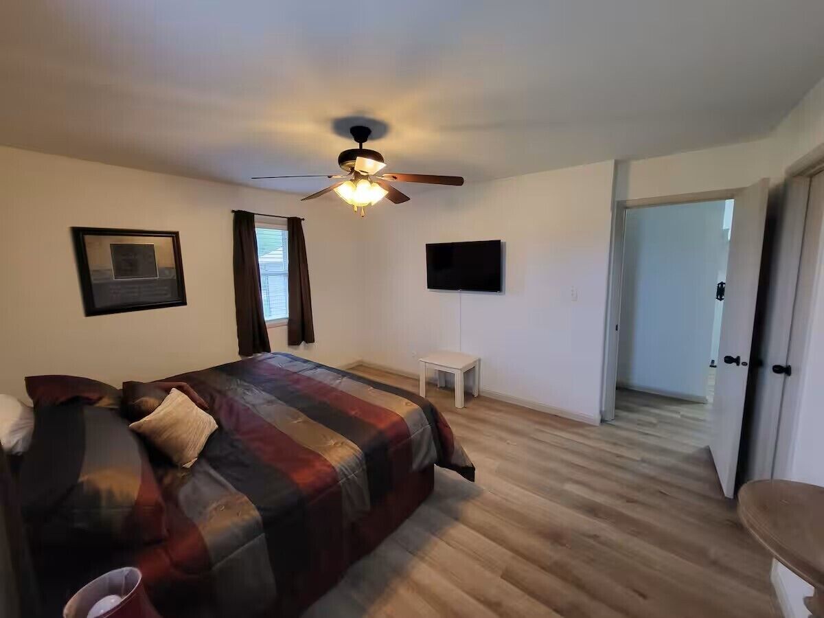 A bedroom with a bed, a ceiling fan, a small table, and a door leading to another room.