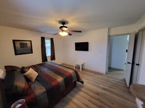 Un dormitorio con cama, ventilador de techo, una mesita y una puerta que da a otra habitación.