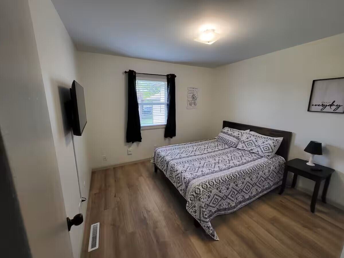 A bedroom with a bed, a nightstand, a wall-mounted TV, a window with curtains, and a poster on the wall.