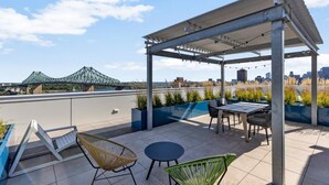 Eine Dachterrasse mit Sitzgelegenheiten im Freien, einer Pergola und Blick auf eine Brücke und die Skyline der Stadt.