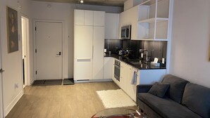 Eine moderne Küche mit weißen Schränken, einer Mikrowelle und einer schwarzen Arbeitsplatte. Im Wohnbereich befindet sich ein graues Sofa und ein rotes Glastisch.