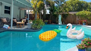 En swimmingpool med oppustelige svaner og ananas-bøjer, omgivet af palmer og et træhegn.