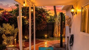 En udendørs terrasse med et glasbord og turkise stole, omgivet af planter og udsigt til solnedgang.