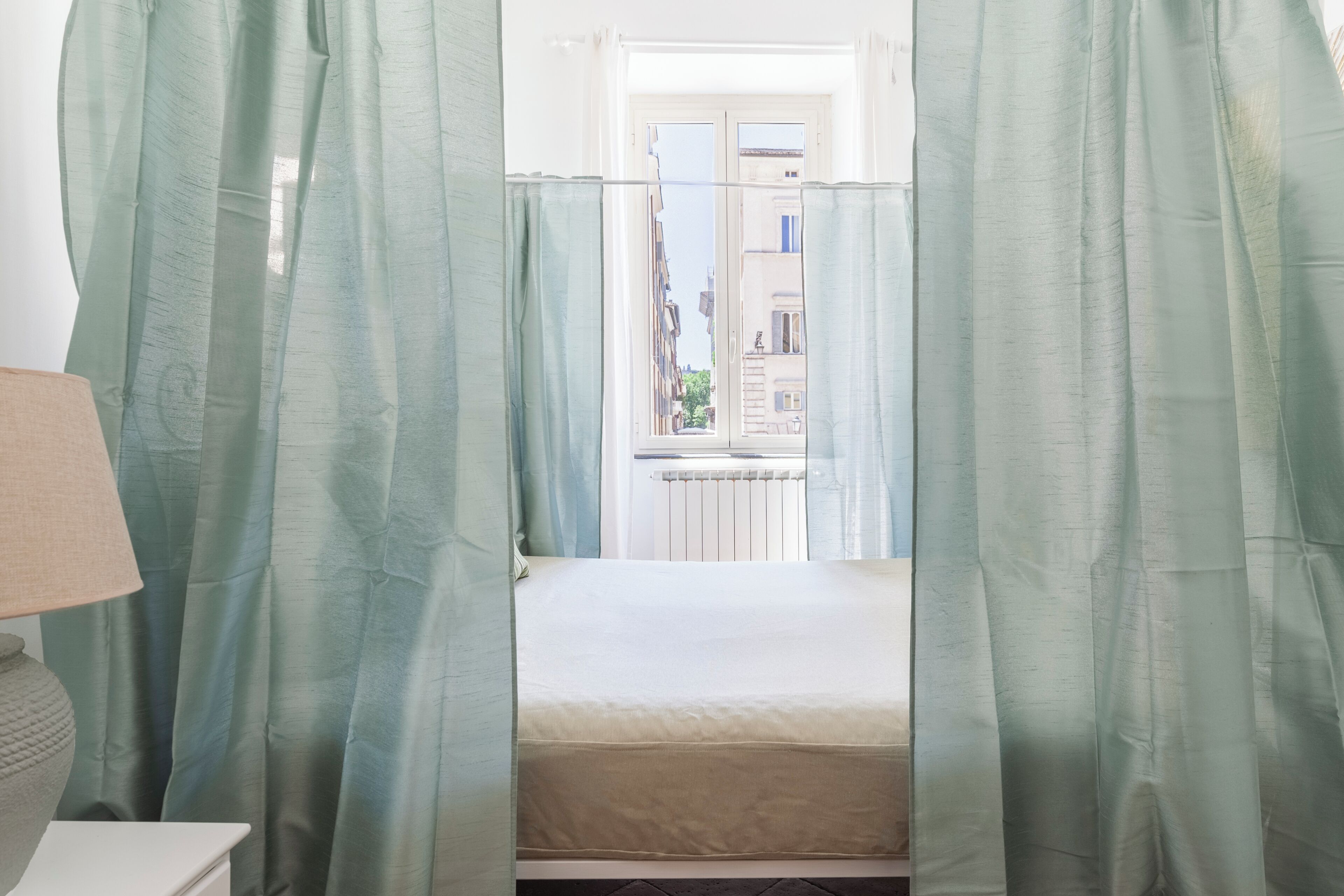 Ein Zimmer mit einem Bett, einem Fenster mit Blick auf Gebäude und einer Lampe auf einem Nachttisch.