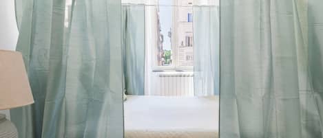 A room with a bed, a window with a view of buildings, and a lamp on a bedside table.
