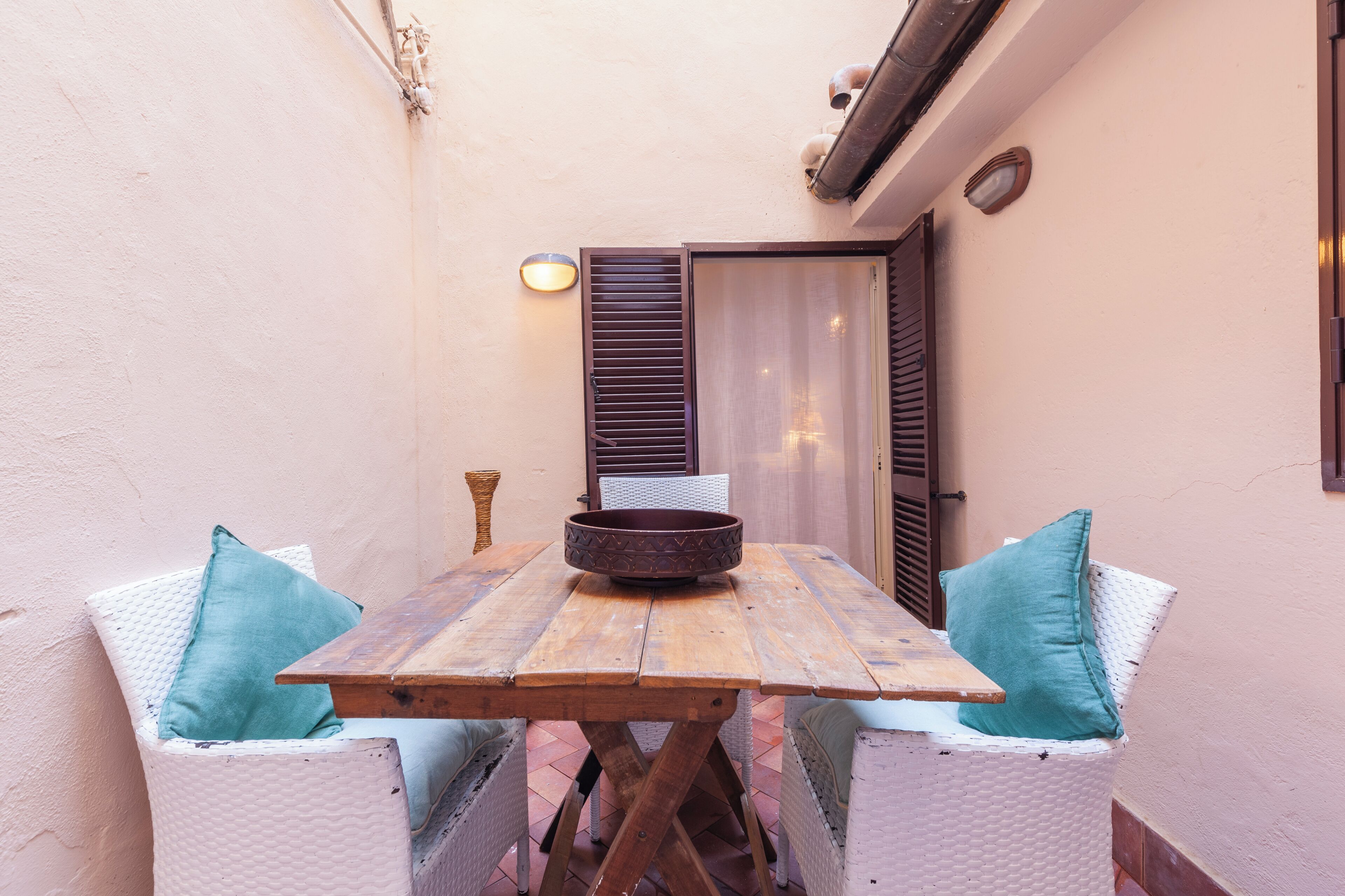 A wooden table with a bowl, surrounded by wicker chairs with teal cushions, in a narrow outdoor space with a door and wall-mounted light fixture.