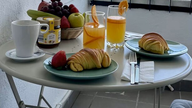 Een tafel met ontbijtartikelen, zoals croissants, fruit en sap, op een balkon met uitzicht op zee.