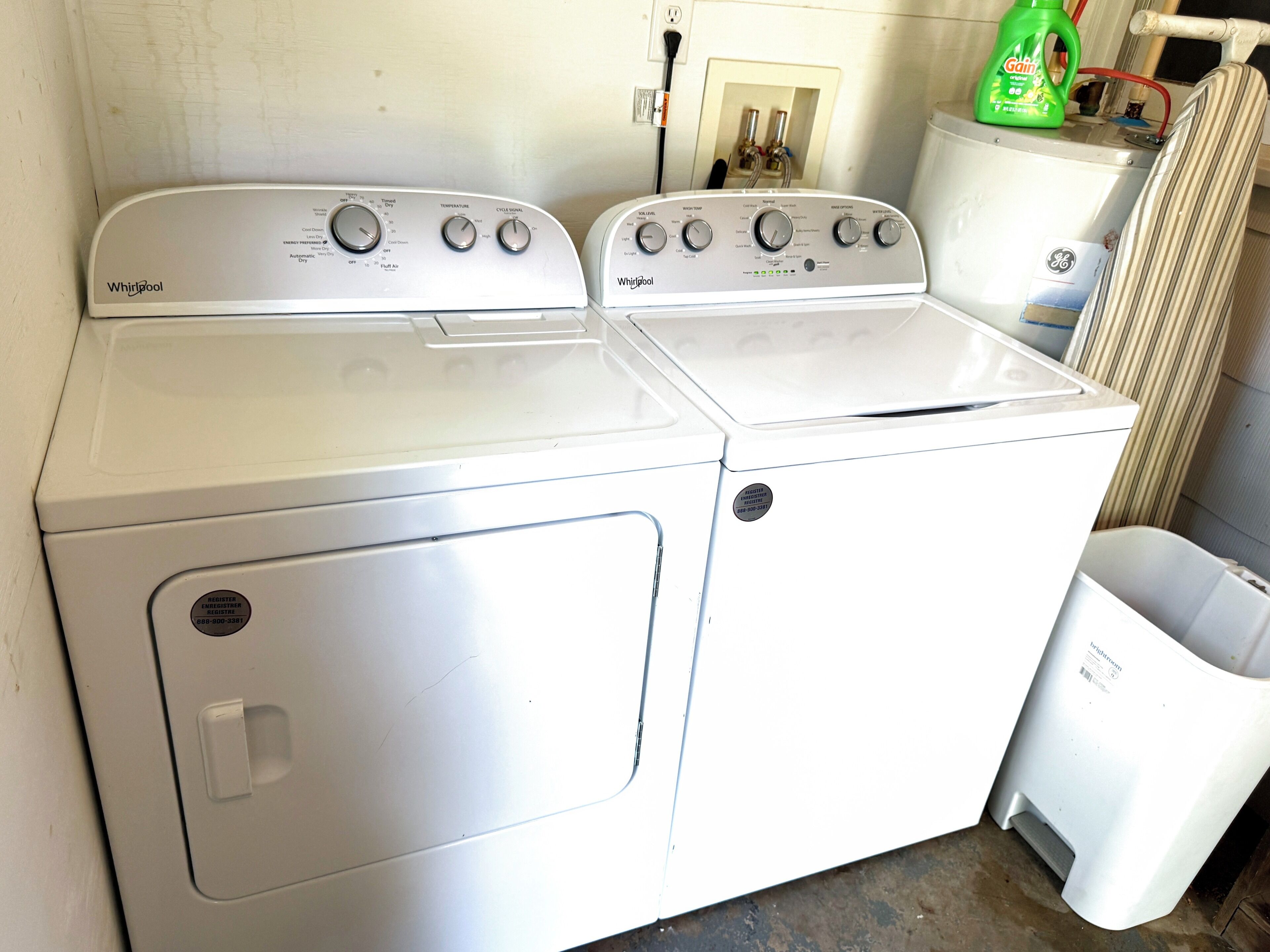 A front view of a Whirlpool washer and dryer unit with control knobs, a power outlet, and a trash can.