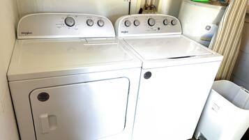 A front view of a Whirlpool washer and dryer unit with control knobs, a power outlet, and a trash can.